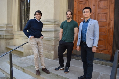 A photo of three men standing outside, at the top of a staircase and in front of an intricately carved set of wooden doors
