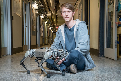 Photo of Ben Katz sitting cross-legged on a hallway floor with a mini-cheetah robot in front of him