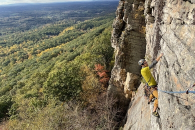Photo of a person rock climbing on cliff with view of mountains and green trees in the distance.