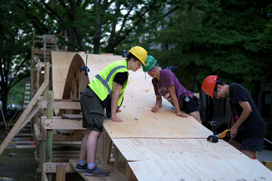 Photo of three students wearing hard hats who are working to build a small wooden roller coaster amid several trees on MIT campus