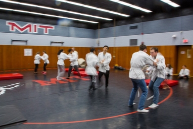 Students practice judo moves one-on-one in a wrestling gym where "M.I.T." is written on the wall