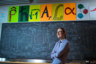 Phiala Shanahan smiles in classroom with blackboard in background, filled with math equations. Colorful posters above of the blackboard show mathematical icons.