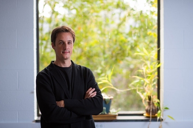 César Terrer stands in front of a window with potted plants on the sill and a green tree outside.