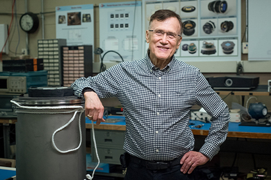 George Ricker stands in a lab, resting his elbow on a large metal cylinder 