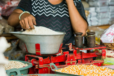 A vendor weighs rice on a red scale in an Indonesian market. Other dried cooking ingredients, including corn and beans are available for sale.