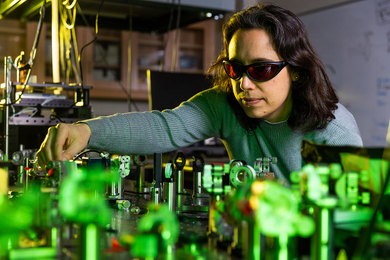 Schlau-Cohen, in a laser lab wearing special glasses, tweaks a device on a table. The room is dark, and the tabletop has green light and many shiny laser parts.