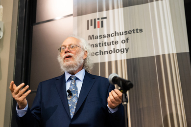 Peter Shor stands at a microphone, speaking, with an MIT banner behind him.