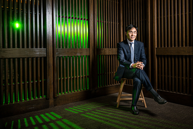 In Song Kim is seated in chair in a room with wooden slatted walls. Through the slats, a green light shines and casts interesting shadows on the ground.