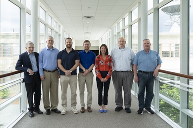 Seven individuals stand side-by-side in a hallway bridge connecting two buildings.