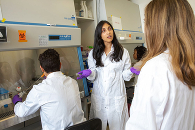 Ritu Raman speaks to two students in a lab. All three are wearing white lab coats and nitrile gloves.