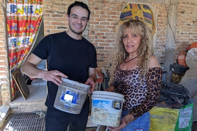 Andres Rico stands next to Eva next to a brick building. Each holds a bucket with a small rectanguar sensor glued to the side.