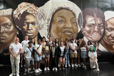 21 people pose in front of a mural depicting faces of five individuals of color