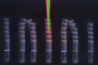 new research in microbiology Seven tall, rectangular columns stand against a black background. The columns are made of a lattice-like pattern and some of them tilt at the top. In the center of the image, a green beam of light reaches between the top of the image and the middle column.