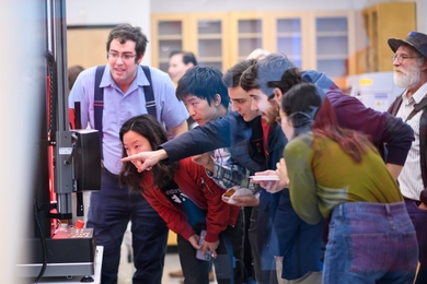 In a lab, six people lean forward to look at something that one of them is pointing at.
