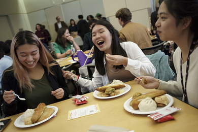 Three students share laughs around a square table while eating food.
