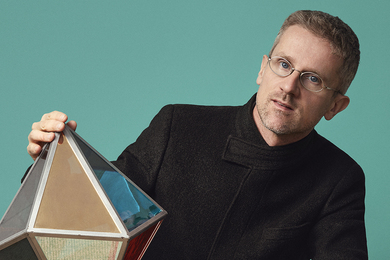 Carlo Ratti poses in a teal room with a geometric figure balanced on his knee