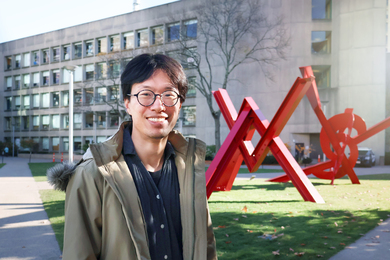 Jonathan Zong poses for a photo on MIT's Hockfield Court, with a cement building and a red sculpture in the background