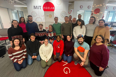 20 members of the AgeLab pose for a group photo in the office, with the red AgeLab logo on the wall and on a rug.