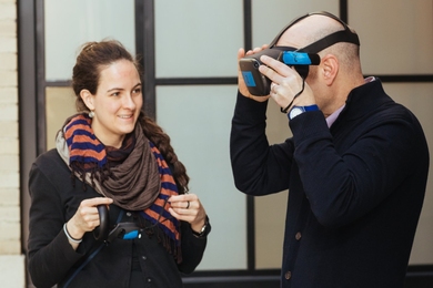 Lydia Brosnahan and an unidentified man stand in front of a window; the man is wearing a virtual reality headset.