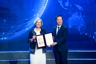 Susan Solomon and Tran Hong Ha pose on a stage. They are both holding a folio open for the audience to see, and Solomon is holding a gold trophy.