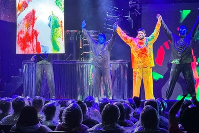A male student clad in a waterproof rain suit covered in multicolored paint stands onstage between two Blue Man Group members with his arms raised, before an audience of people wearing clear ponchos.