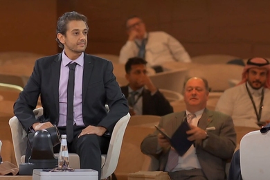 Mohammed Yahia sits on a stage. He's wearing a microphone and speaking to someone out of the frame. An out-of-focus audience is in the background. 