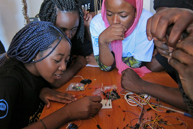 Three African children lean over a table, working on a circuit board, with more people outside the frame