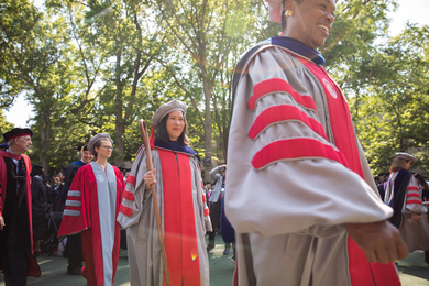 Lily Tsai, along with MIT administration and faculty, proceed through Killian Court on a spring day. All are wearing academic regalia and Tsai holds a wooden staff.