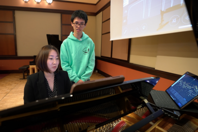 Mi-Eun Kim, seated, plays a piano while Holden Mui, standing behind her, watches. An open laptop with a visual representation of data rests atop the piano.