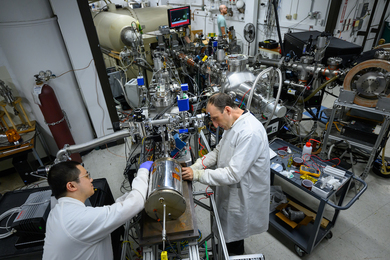 Weiyue Zhou and Michael Short, both in white lab coats, attach a silver cylinder the size of a small wastecan to the end of a proton accelerator, a large room-sized machine with many connecting tubes and wires. 