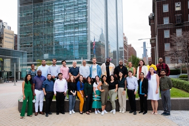 28 people stand in two rows, smiling, on a city block with a large glass building in the background