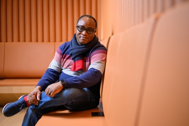 Ericmoore Jossou, a Black man with dark-rimmed glasses, sits on a orange leather-covered corner bench in a room lined with the same color walls.