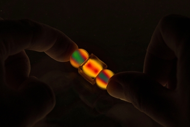 In darkness, thumbs hold two small spherical and one small cube objects that light up in vivid yellow, red, and green.