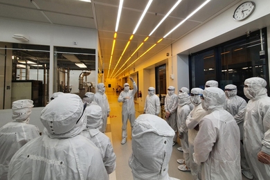 A view from behind of about 15 people dressed head-to-toe in white cleanroom suits, facing another gowned-up individual gesturing as they speak.