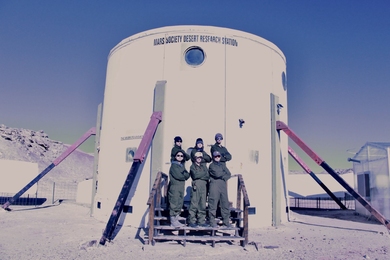 Six people in jumpsuits stand with their arms folded in front of a two-storey cylindrical structure labeled "Mars Society Desert Research Station" on a cloudless day.