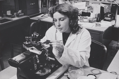 Grayscale photo of Anne Serby wearing a lab coat and seated at a lab table using a microscope