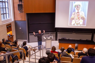 Master Bladesmith Bob Kramer speaks in front of an audience in a classroom with stadium seating. Behind him on a screen is a picture of Kramer as a circus clown.