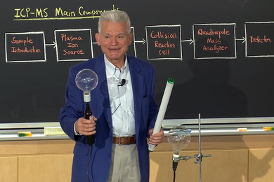 Male instructor teaches in front of a blackboard that contains a series of terms including "plasma ion source," and "collision reaction cell."  He is holding a small round lightbulb in one hand and a long fluorescent lightbulb in the other.