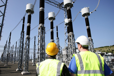 Two men in hardhats and safety vests, seen from behind, inspect a forest of electrical pylons and wires on a cloudless day