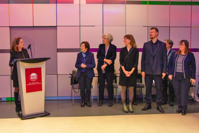 At left, Cynthia Barnhart speaks at a podium while about half a dozen indiviuals look on. The event is in a room lined with white square tiles and colored accent tiles.