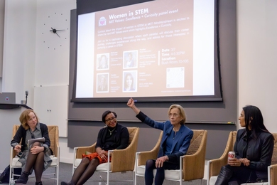 Four women sit on a stage, one with a raised fist, in front of a projected slide headlined "Women in STEM." 