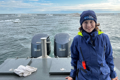 Emma Bullock smiles while near the back of a boat and wearing waterproof gear, with the ocean and sky in background.