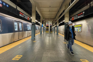 A man wearing a protective masks walks down an empty New York subway station, with silver subways cars on the left and right sides.