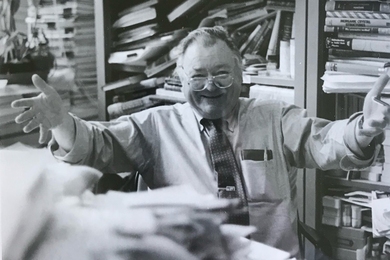 A grayscale photograph of Professor Bernie Wuensch in his office, surrounded by books and heaps of papers, welcoming the camera with open arms and a warm smile