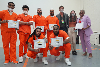 In a white room, six individuals in orange jumpsuits are smiling and holding certificates of achievement. They are accompanied by three individuals dressed in casual and professional attire.