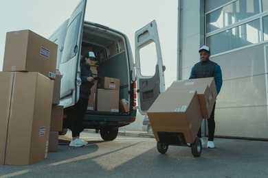 A man moves three large boxes on a handtruck while a woman standing in back of an open van takes inventory