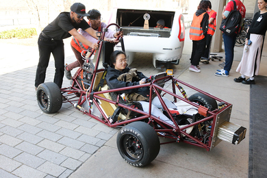 Two students push the tubular steel Motorsports car into Lobby 13 while a third sits in the car and steers