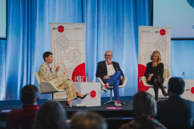 Three people sit on a stage, one of them speaking. Red and white panels with the MIT AgeLab logo are behind them.