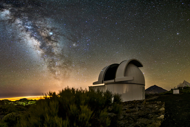 A night-time photo shows a white, cylindrical observatory with the roof open part-way, in a rural landscape. The sky is full of the Milky Way and stars.