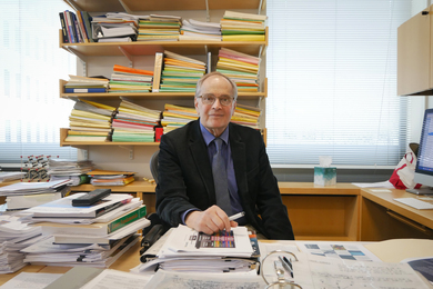 John Joannopoulos sits in his office full of stacks of paper, binders, and folders.